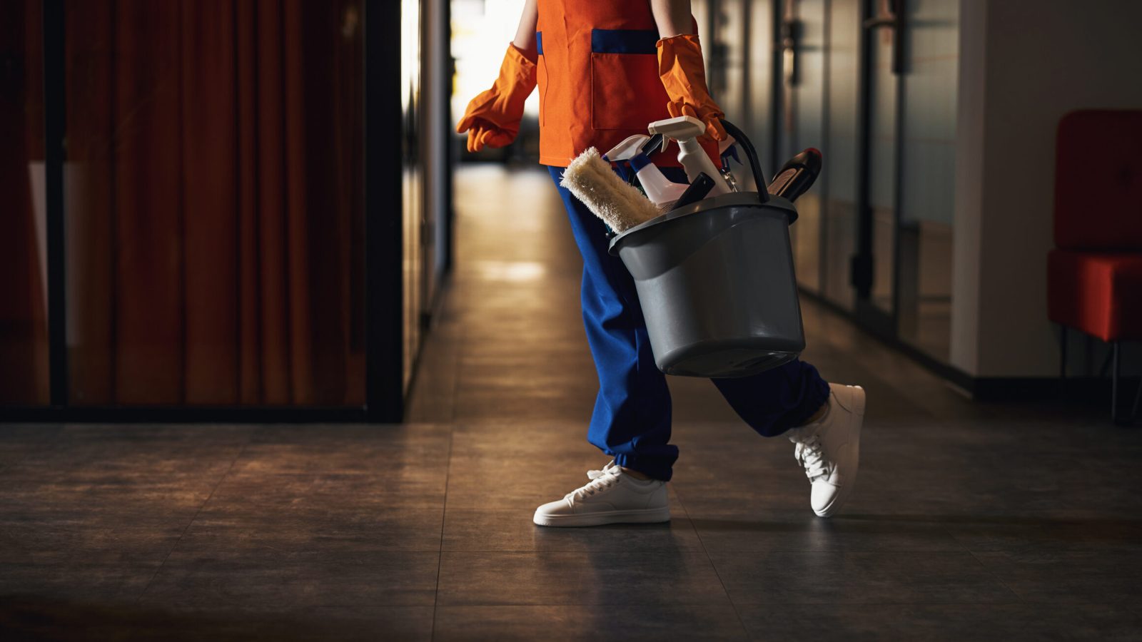 Cleaner in uniform carrying a bucket of janitorial supplies while at work.